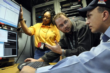 Civilian employees work with Information Systems Technician 2nd Class James Rago to troubleshoot the video teleconference system of a video information exchange system.