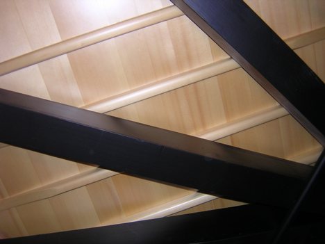 View from below of a 182-cm grand piano. In order of distance from viewer: softwood braces, tapered soundboard ribs, soundboard. The metal rod at lower right is a humidity control device.