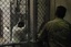 A Sailor assigned to the Navy Expeditionary Guard Battalion stands watch over a cell block in Camp 6 at Joint Task Force (JTF) Guantanamo while detainees look through magazines and books.