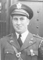 ead and torso of a smirking white man wearing a peaked cap and a military jacket, adorned with pins and badges and a strap running diagonally across his chest, over a shirt and tie.