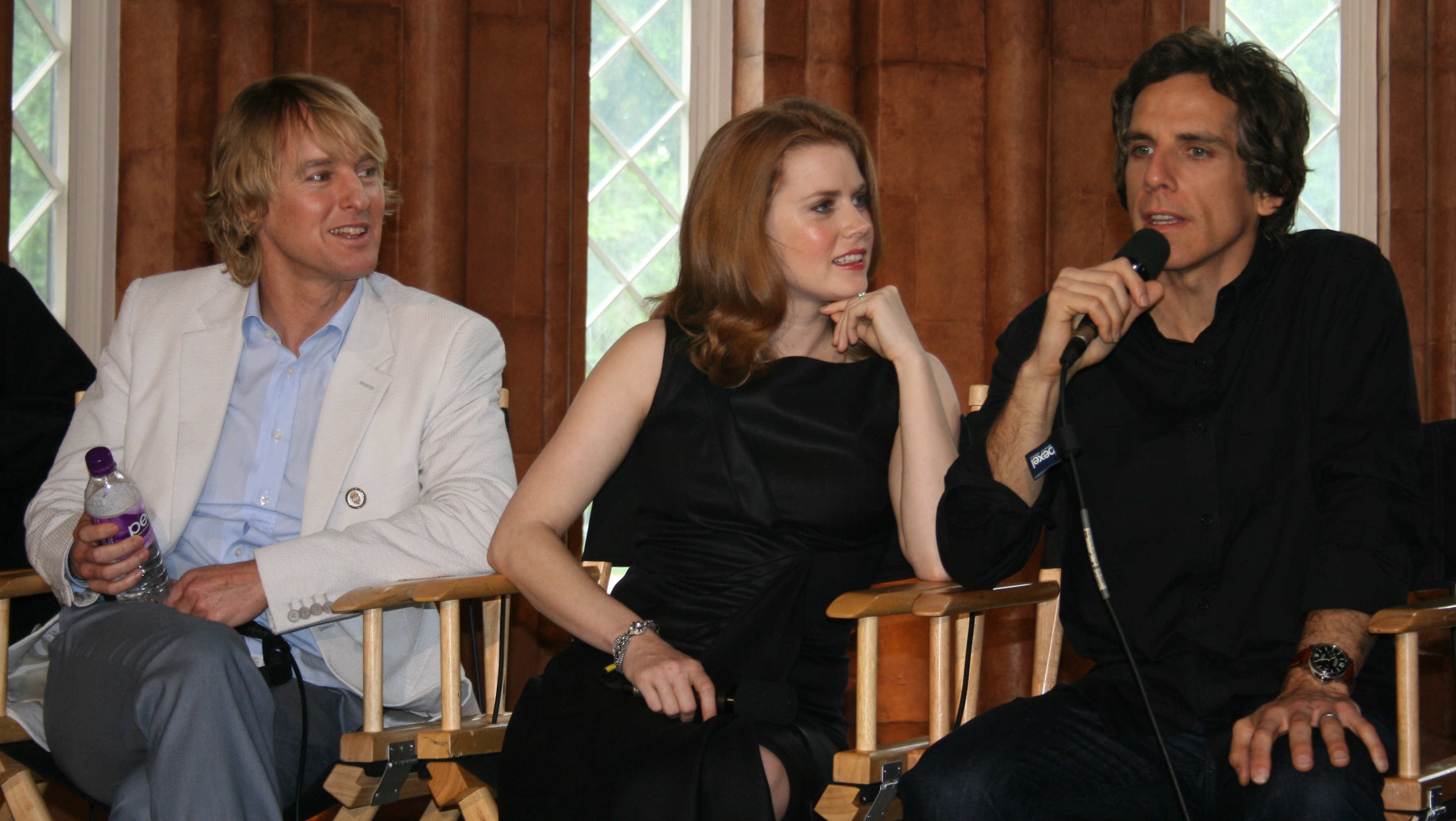 Casual photo of a young woman sitting between two men in director chairs. The woman is wearing a black, sleeveless dress and is intently listening to one of the men, Ben Stiller, talk into a microphone.