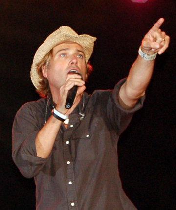 Michael W. Smith in a brown western-style shirt wearing a straw cowboy hat, on stage during a performance. He is holding a cordless microphone in his right hand and pointing into the audience with his left.