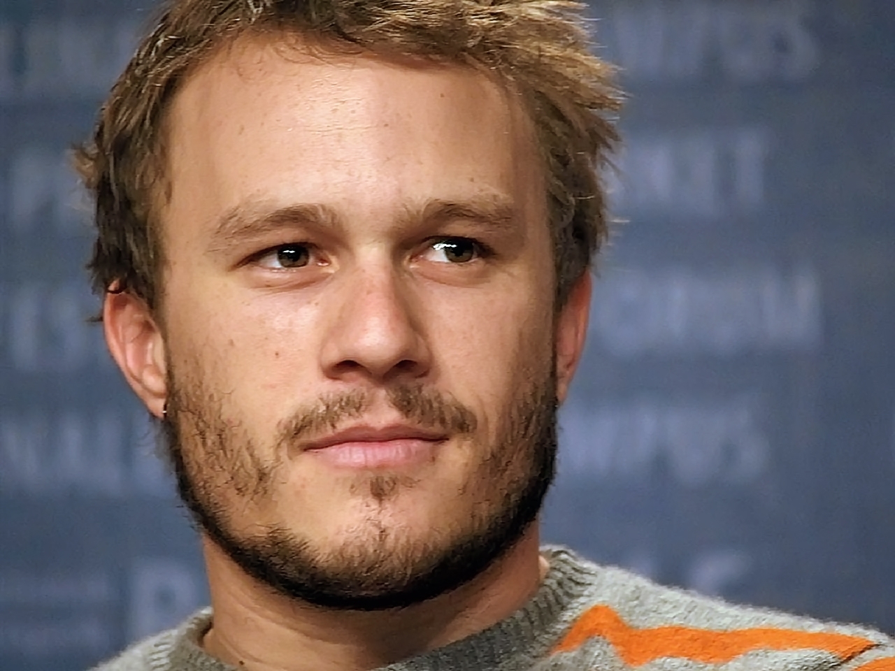 Close up of a man's face with brown eyes, tousled brown hair and scraggly beard growth. He is looking toward his left. He is wearing a grey jumper with an orange stripe near his left shoulder and upper left arm. The background is blue with out of focus writing.