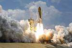 Sandwiched by billowing clouds above and smoke and steam clouds below, space shuttle Atlantis hurtles past the lightning mast on top of the fixed service structure on Launch Pad 39A at NASA's Kennedy Space Center in Florida.