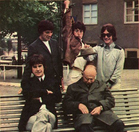 On a bench in a park sit five men, two seated and three standing behind the bench. Clockwise from left is a man in a black suit with Khaki pants; a man in a black suit with black pants; a man wearing a brown coat with Khaki pants and raising his arm jokingly as if to stab the sleeping man below him; a man, wearing a grey/light green coat and Khaki pants and holding out a hat above the sleeping man's head. The sleeping man is wearing green.