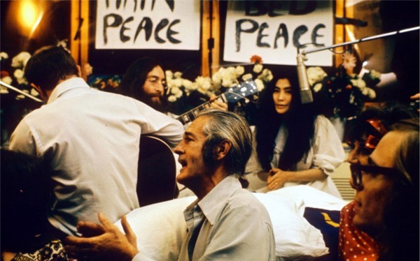 Lennon and Ono sit in front of flowers and placards bearing the word "peace." Lennon is only partly visible, and he holds an acoustic guitar. Ono wears a white dress, and there is a hanging microphone in front of her. In the foreground of the image are three men, one of them a guitarist facing away, and a woman.