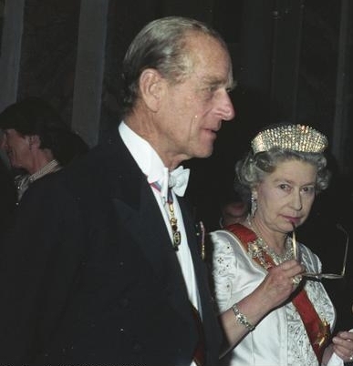 Behind her husband, Elizabeth holds a pair of spectacles to her mouth in a thoughtful pose