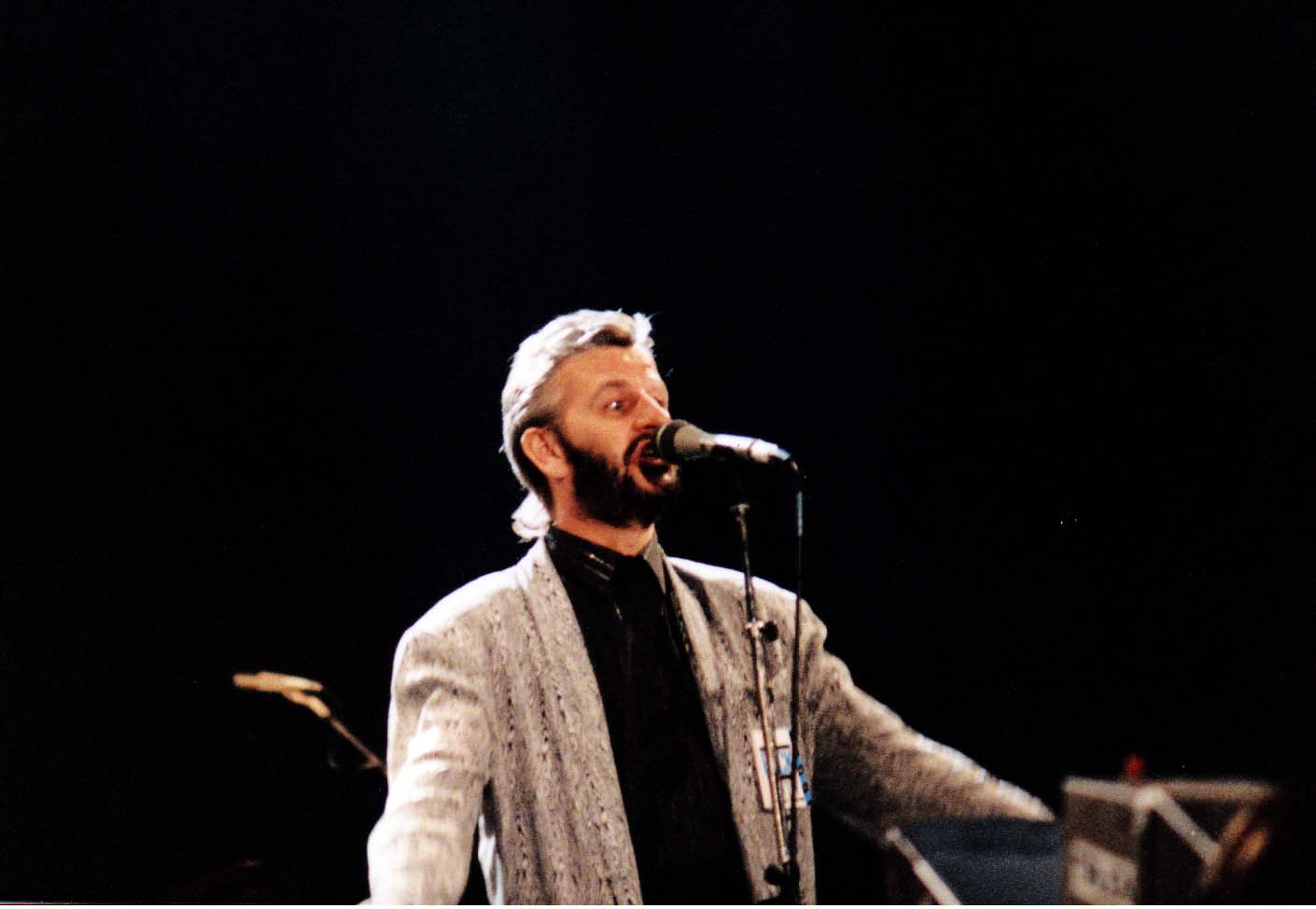 Starr in his forties, wearing a grey jacket and a black shirt, standing behind a microphone and singing.