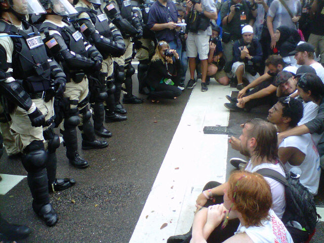 (A photograph of a line of police in body armor and riot gear standing in front of a group of seated, unarmed protesters in casual dress.)
