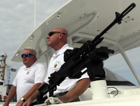 (A photograph of two police officers on a boat with a mounted carbine pointing off toward the top of the photo.)