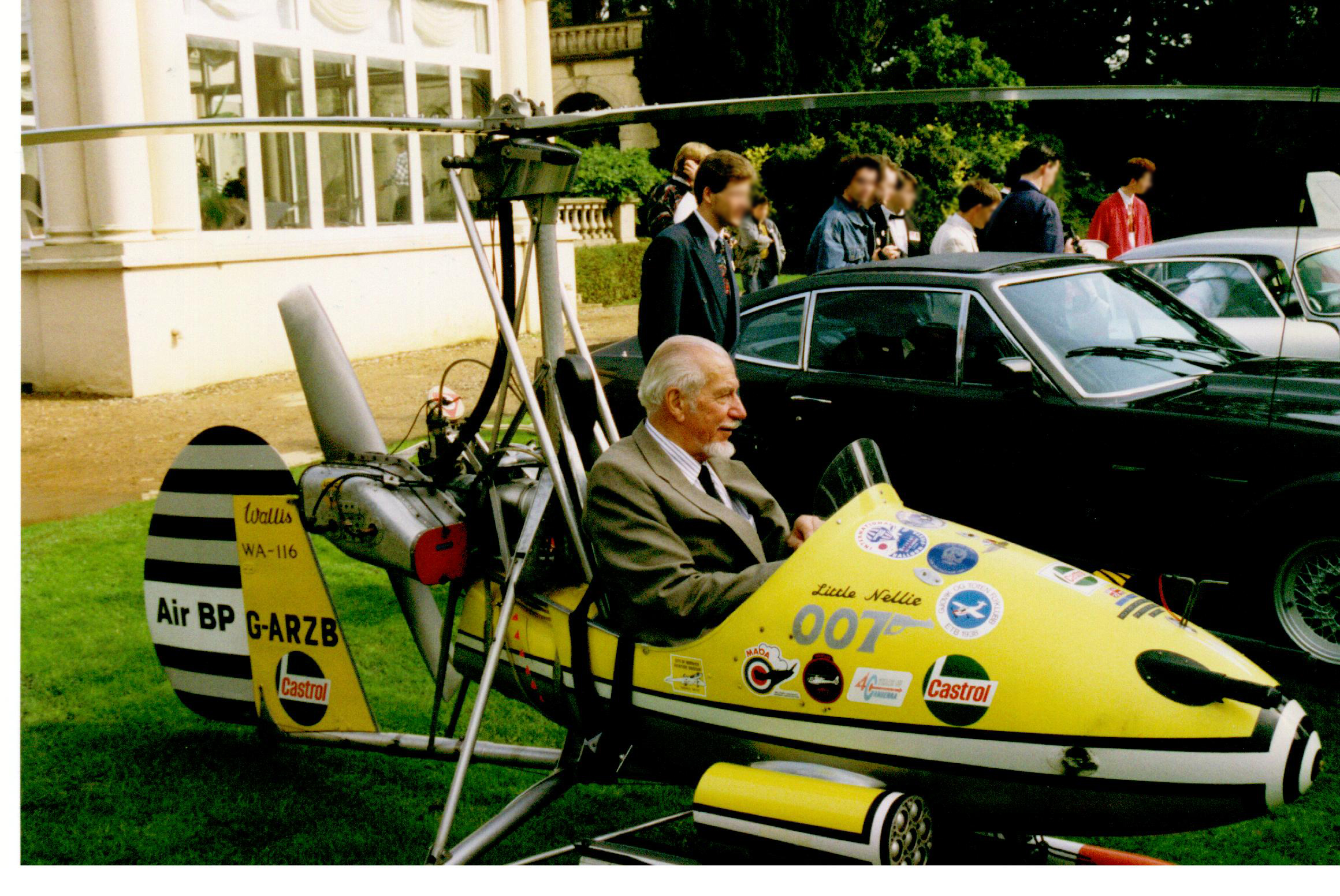 Small, one man, open-cockpit helicopter on a lawn about the size of a car next to it, with a man sitting in it.