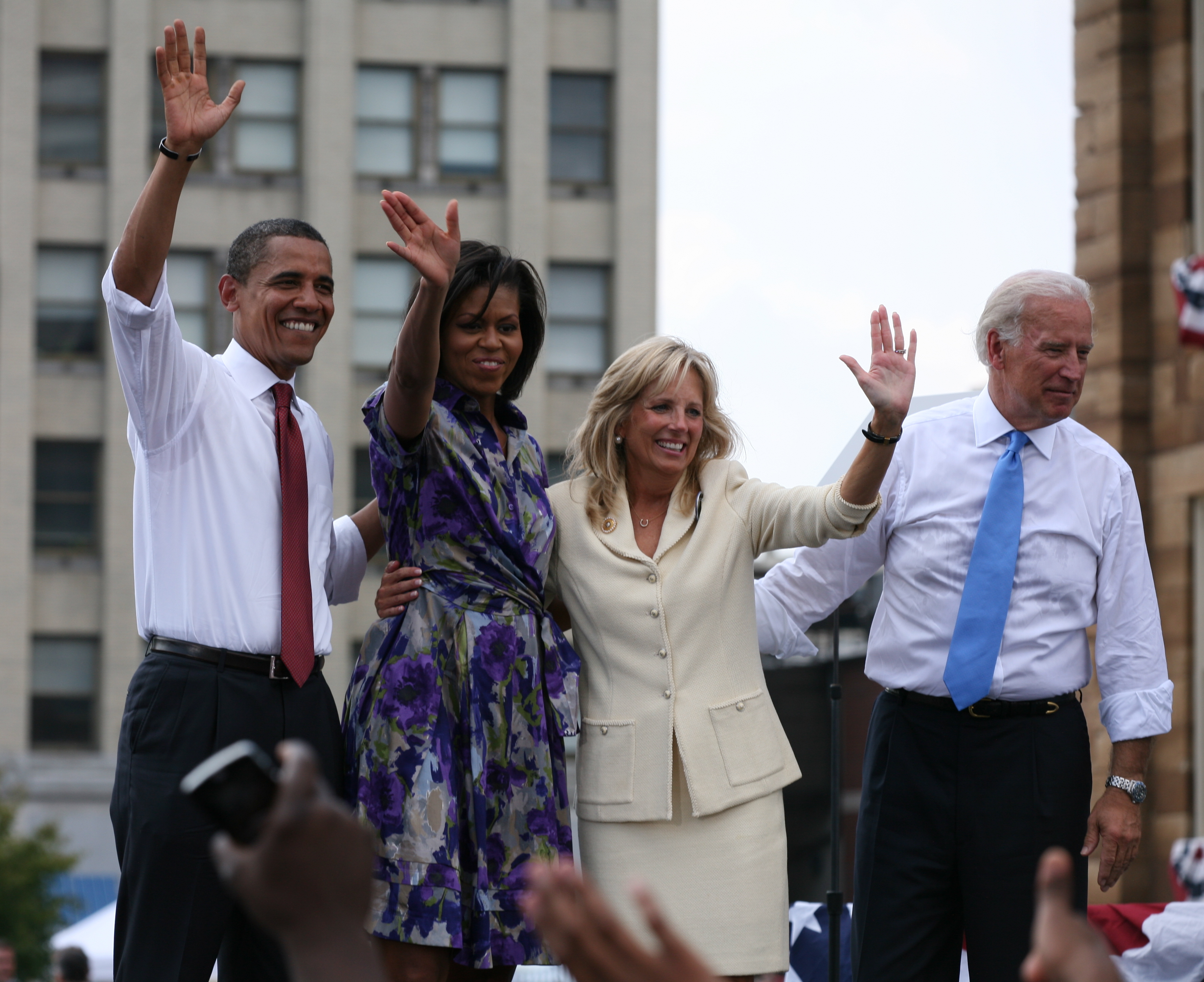 Barack and Michelle Obama and a woman and a man on an outdoor stage. The first three smile and wave. The men wear suit pants, white shirts with the sleeves rolled up, and ties. Michelle is in a colorful print dress and the other woman is in a creme business suit.