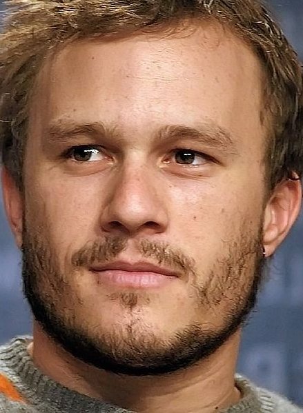 Close up of a man's face with brown eyes, tousled brown hair and scraggly beard growth. He is looking toward his left. He is wearing a grey jumper with an orange stripe near his left shoulder and upper left arm. The background is blue with out of focus writing.