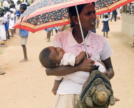 File - A mother carrying her child awaits the arrival of Secretary-General Ban Ki-moon at the Manik Farm camp of the Internally Displaced Persons (IDPs), Sri Lanka, 23 May, 2009.