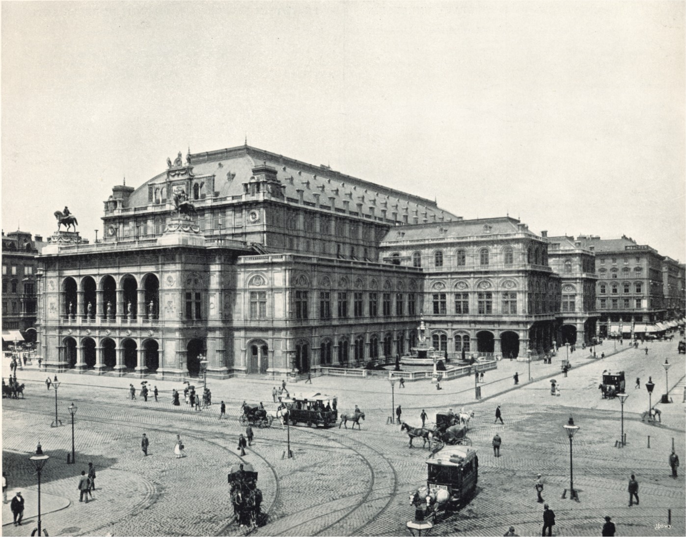  An imposing, heavily ornamented building in a city location, with numerous horsedrawn vehicles and pedestrians passing. There are visible tramlines in the street.