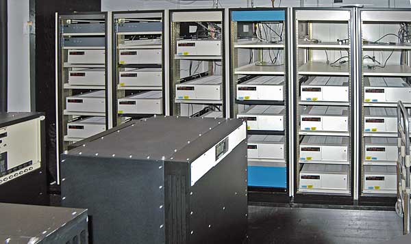  A room with a black box in the foreground and six control cabinets with space for five to six racks each. Most, but not all, of the cabinets are filled with white boxes.