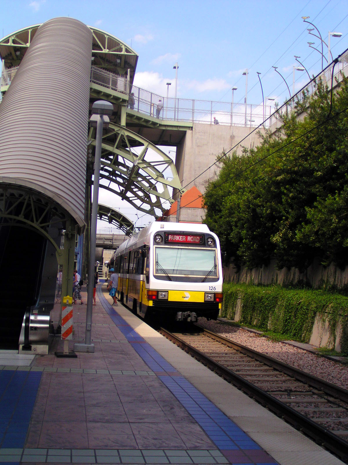 An escalator descends from the street to an island platform station with a white and yellow train present along a landscaped track.