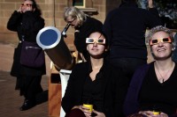 Women wearing protective glasses sit near another woman looking through a telescope at the annular solar eclipse from atop Observatory Hill in Sydney