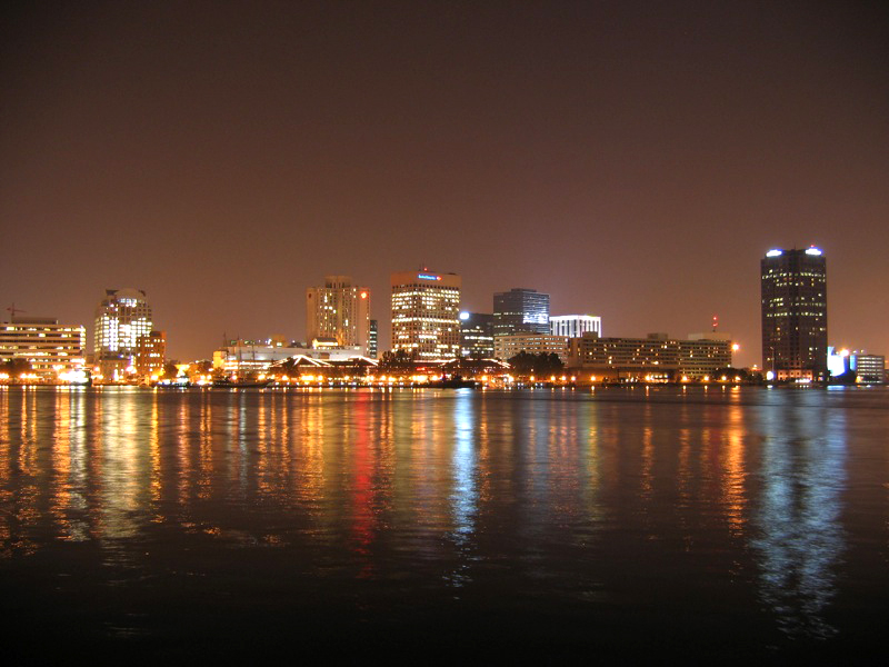 Nighttime panorama featuring lights from skyscrapers reflecting off black water in shades of red, blue, and yellow.