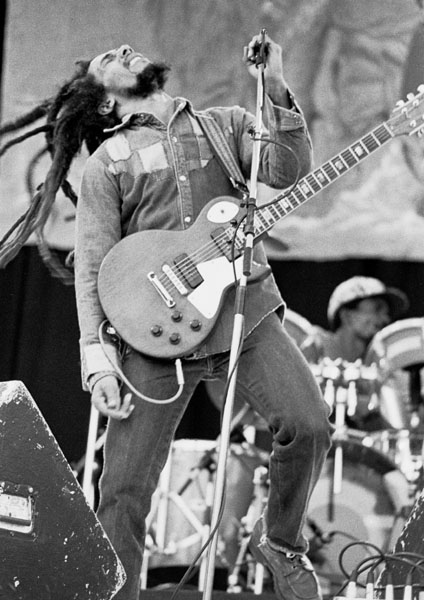 Black and white picture of a man with long dreadlocks playing the guitar on stage.