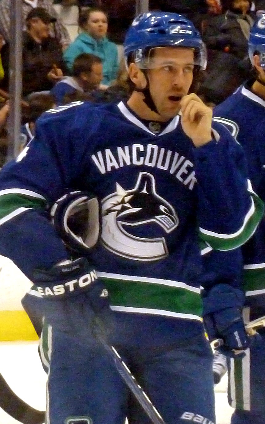 A Caucasian ice hockey player. He is looking to the right with his hand at his mouth. He wears a blue, visored helmet and a blue jersey with white and green trim.