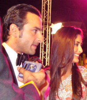 A man and woman facing to the left. The man has black hair and sharp features, while wearing a tuxedo. Somebody is holding a tape recorder in front of his mouth. The woman wears a white sari and has flowing brown hair. She is smiling.