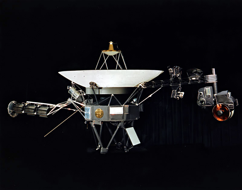 A space probe with squat cylindrical body topped by a large parabolic radio antenna dish pointing upwards, a three-element radioisotope thermoelectric generator on a boom extending left, and scientific instruments on a boom extending right. A golden disk is fixed to the body.