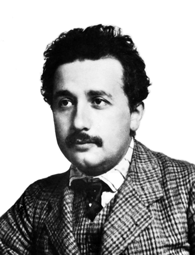 Head and shoulders shot of a young, moustached man with dark, curly hair wearing a plaid suit and vest, striped shirt, and a dark tie.