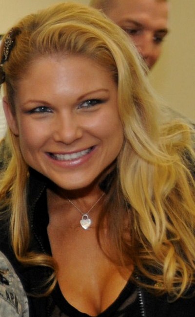 A blond woman smiles while wearing a black top, a silver necklace and a brooch in her hair.