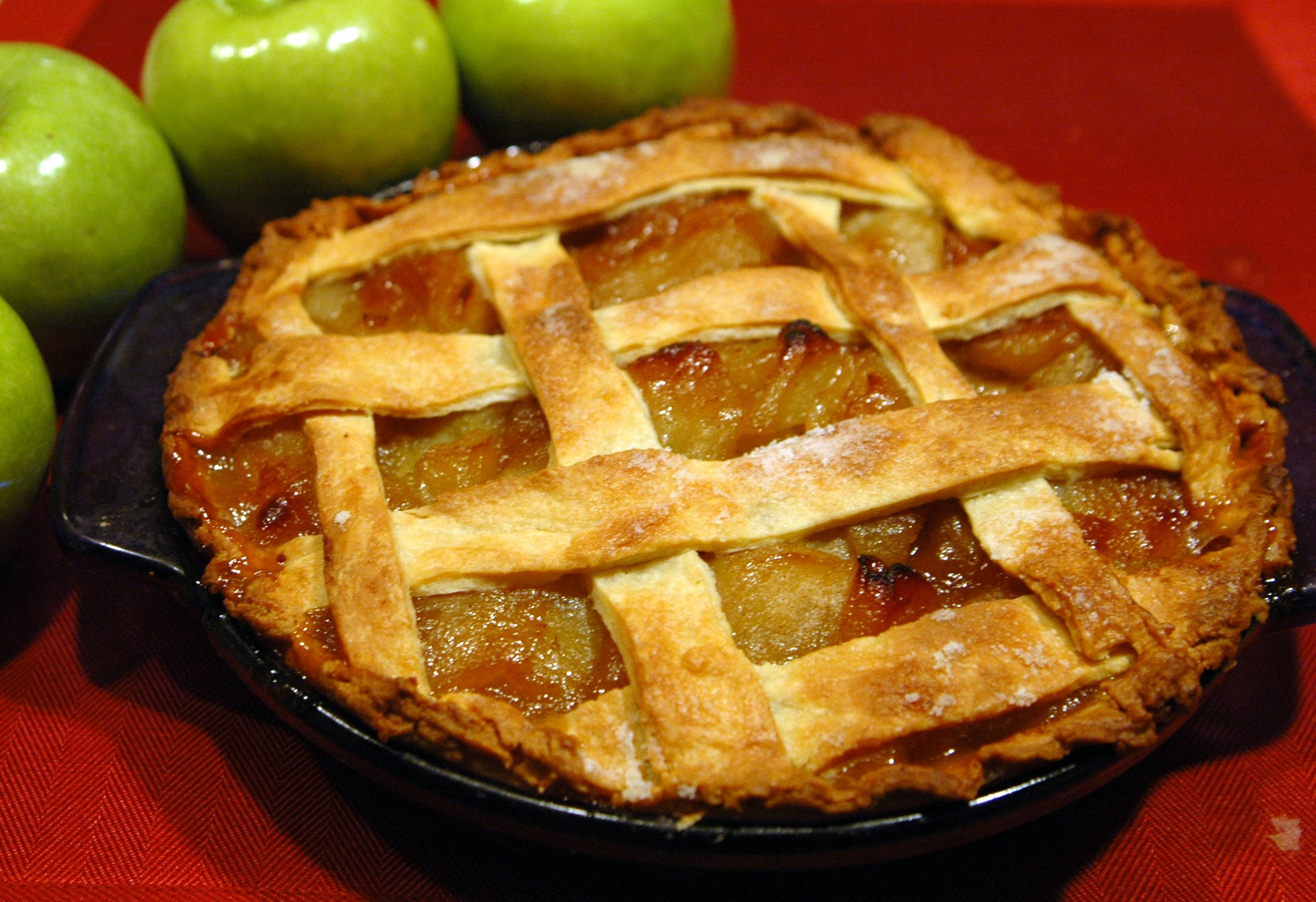 An apple pie on a red table cloth, with green apples next to it.