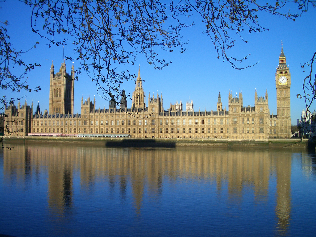 Photograph of rectangular floodlight building, reflected in water. The building has multiple towers including one at each end. The tower on the right includes an illuminated clock face.