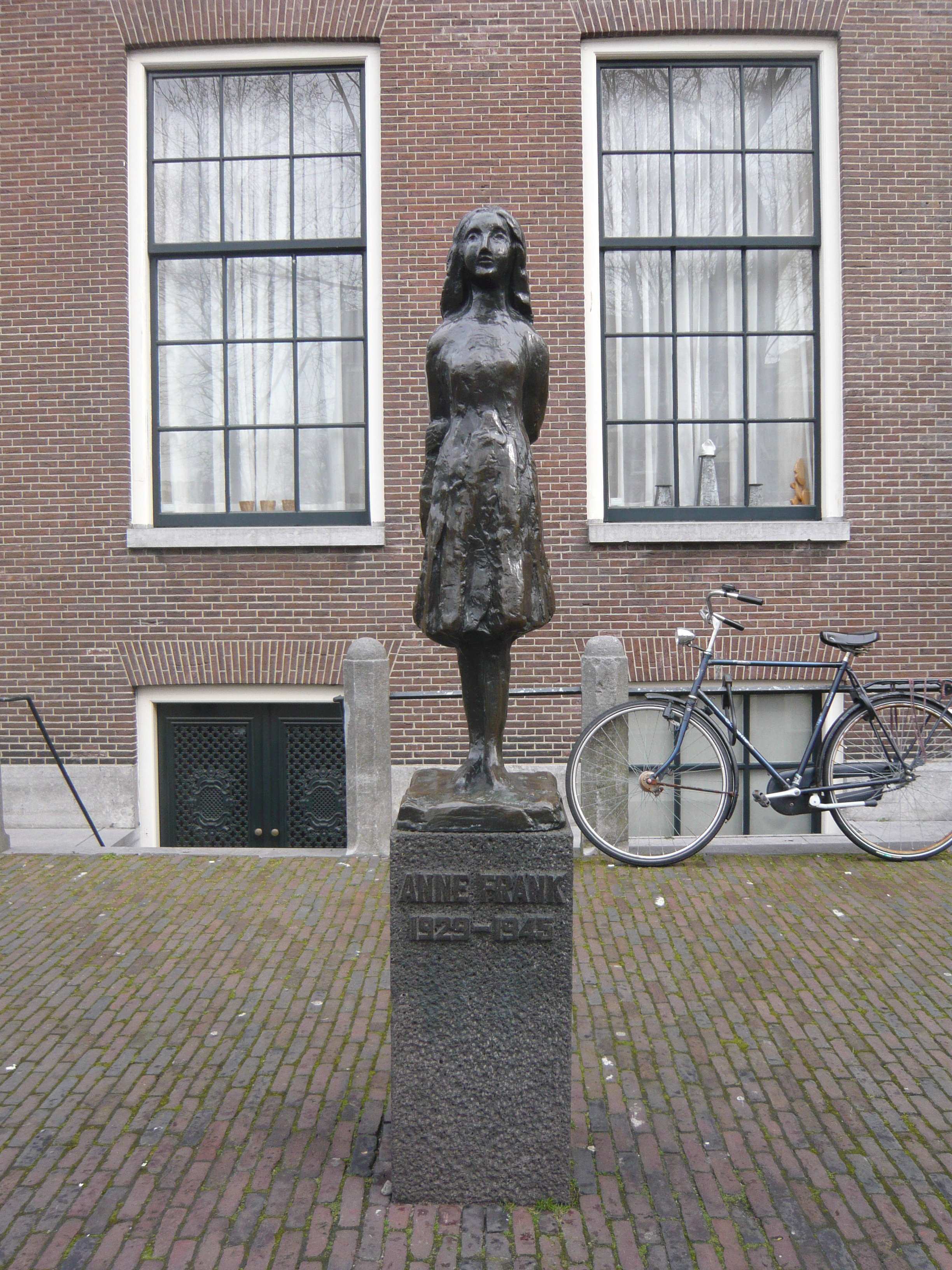 A bronze statue of a smiling Anne Frank, wearing a short dress and standing with her arms behind her back, sits upon a stone plinth with a plaquereading "Anne Frank 1929–1945". The statue is in a small square, and behind it is a brick building with two large windows, and a bicycle. The statue stands between the two windows.