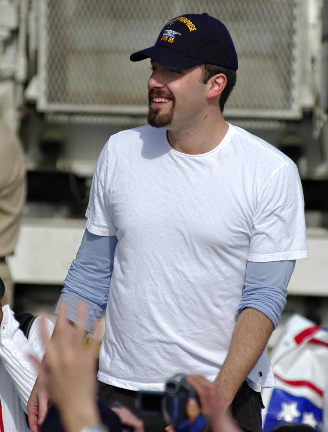 Smiling young man with a trim goatee and moustache, wearing a white t-shirt and a baseball cap. He is surrounding by hands reaching out to him.