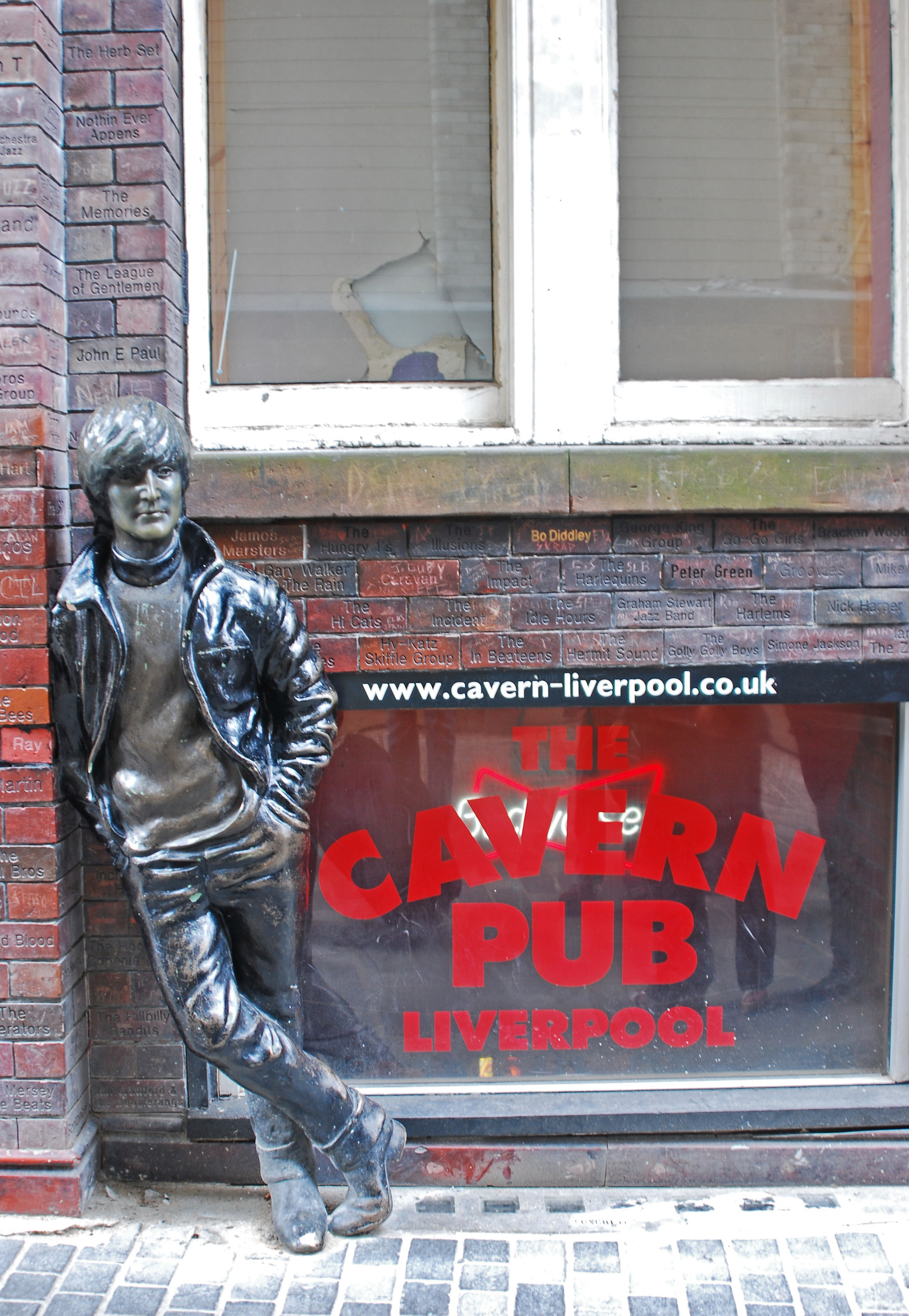 A statue depicting a young Lennon outside a brick building. Next to the statue are three windows, with two side-by-side above the lower, which bears signage advertising the Cavern pub.