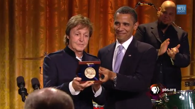  Mcartney being handed an award from President Obama, both men are wearing dark suits, an orange curtain can be seen behind them, as well as drummer Abe Laboriel Jr., who is clapping