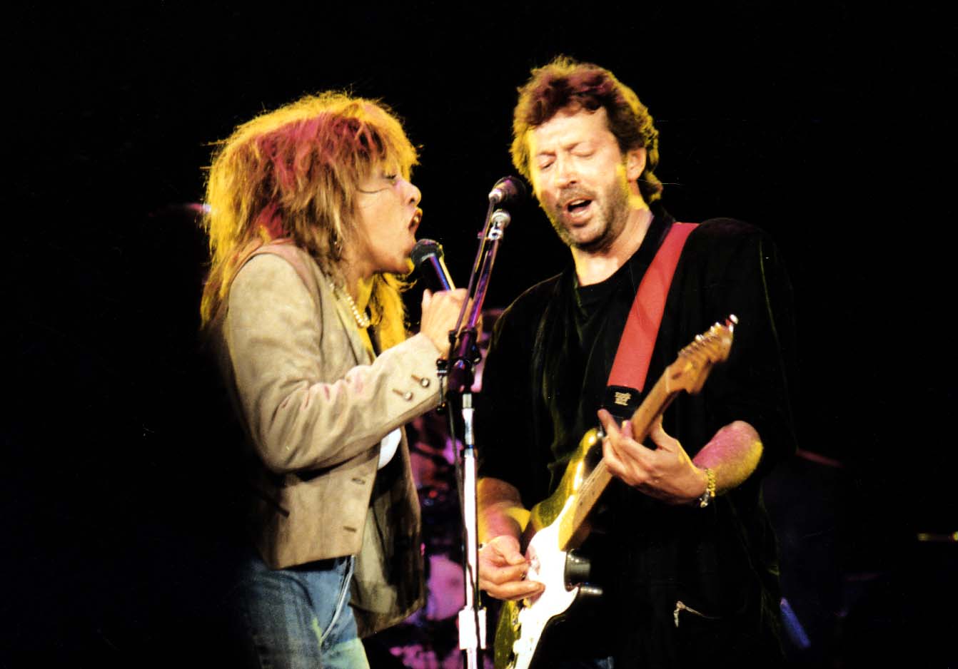 Turner and Clapton, on stage, sharing a microphone stand, singing.