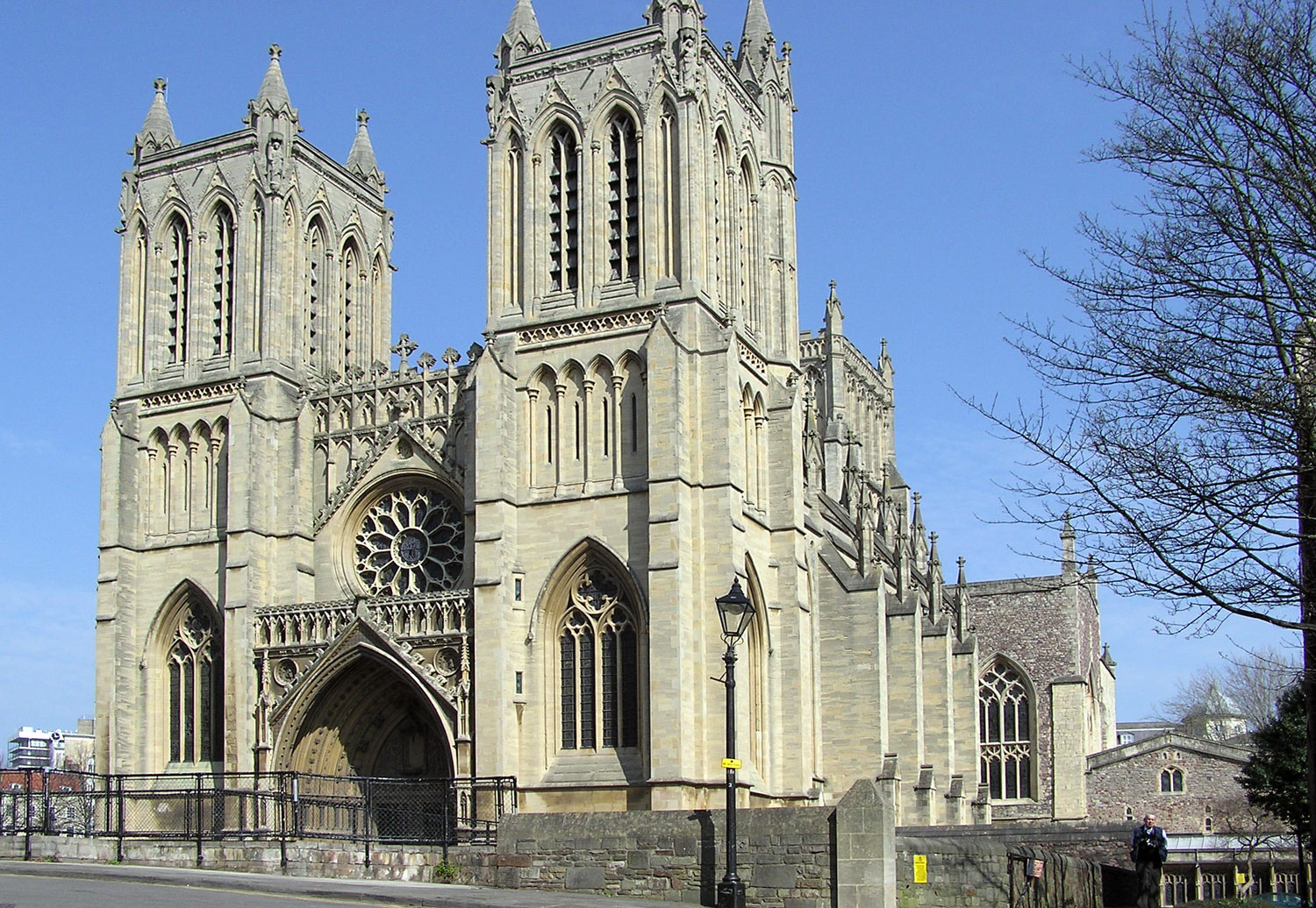 A stone built Victorian Gothic building with two square towers and a central arched entrance underneath a circular ornate window. A Victorian street lamp stands in front of the building and on the right part of a leafless tree, with blues skies behind.