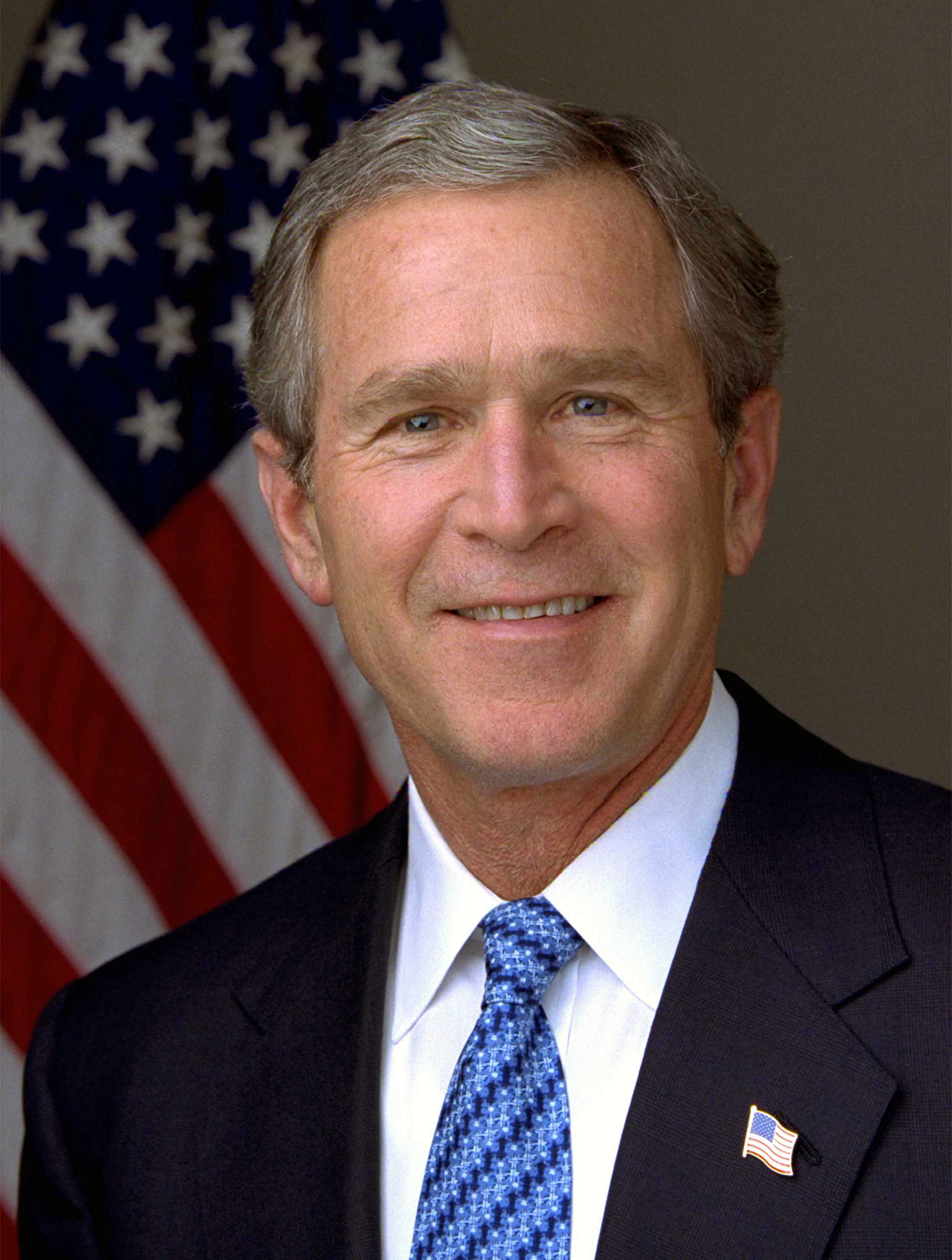 A portrait shot of a smiling older male looking straight ahead. He has short gray hair, and is wearing a dark navy blazer with a blue styled tie over a white collared shirt. In the background is an American flag hanging from a flagpole.