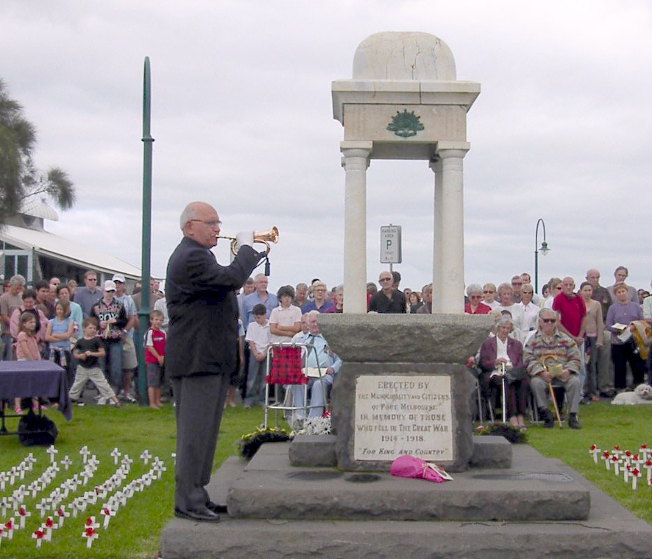 A balding man wearing a suit and playing a bugle, while standing in front of a crowd of other people and a stone monument.