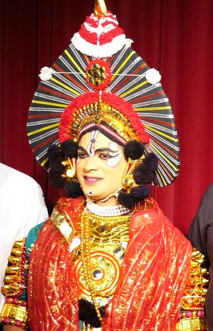 Person with painted eyes in Yakshagana costume, as gold-spangled robe with red sheer scarf and spiked headress on gold crown