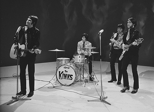 Four members of the band The Kinks stand onstage, during a television appearance. From left to right is Ray Davies, wearing a leather suit and long black pants. He is strumming an acoustic guitar and preparing to sing into a microphone placed in front of him. Next is Mick Avory, seated and playing the drums. He is wearing a bright colored long sleeve shirt and dark pants. After him is bassist Pete Quaife, playing his instrument and wearing set of clothes similar to Avory's. Last is Dave Davies, wearing what appears to be a leather suit with a short tie, black pants and high-heeled loafers. He plays an elaborate v-shaped electric guitar and is standing behind a microphone.