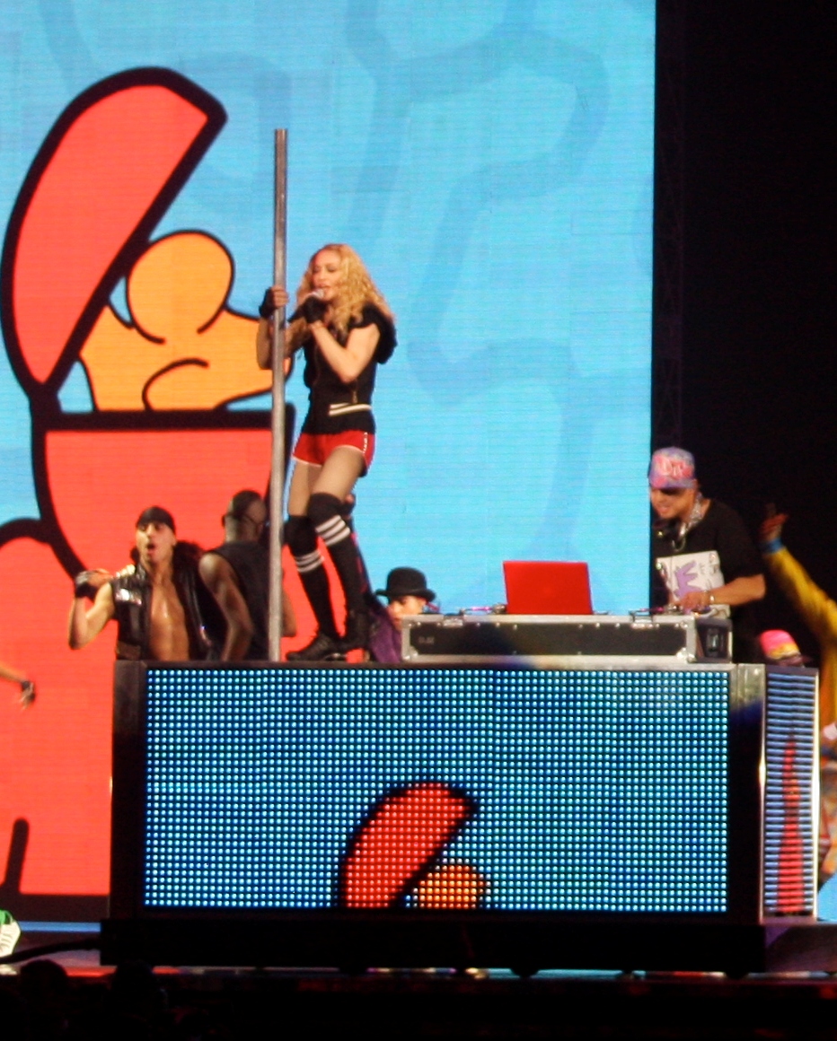 A blond woman stands atop a rectangular black box. She wears red shorts and sings into a microphone, while holding a pole atop the box, with her right hand. A man is seen beside her, wearing a cap. The backdrop behind them display light blue patterns.