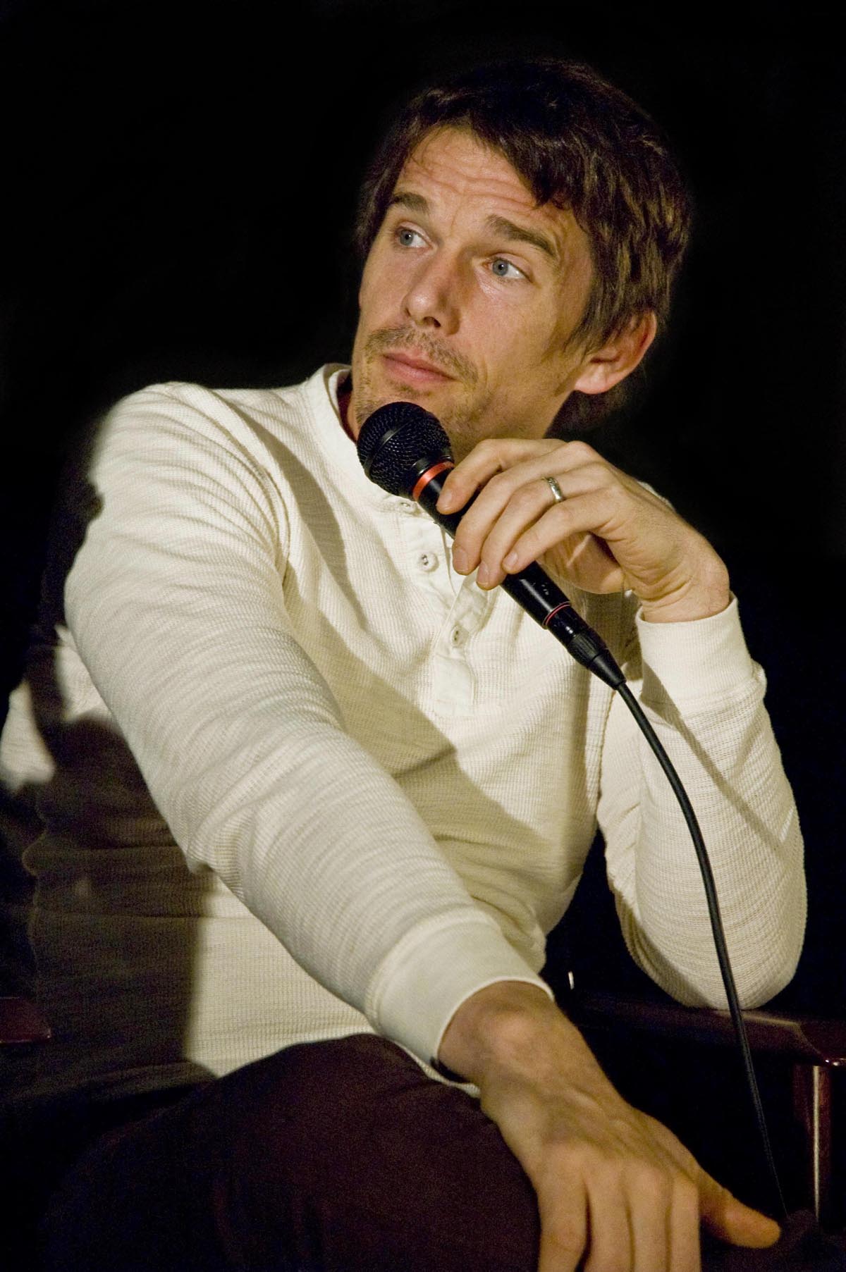 A Caucasian male with light brown hair and light brown stubble is facing to the left. He is wearing a white long sleeve shirt, and is holding a black microphone.