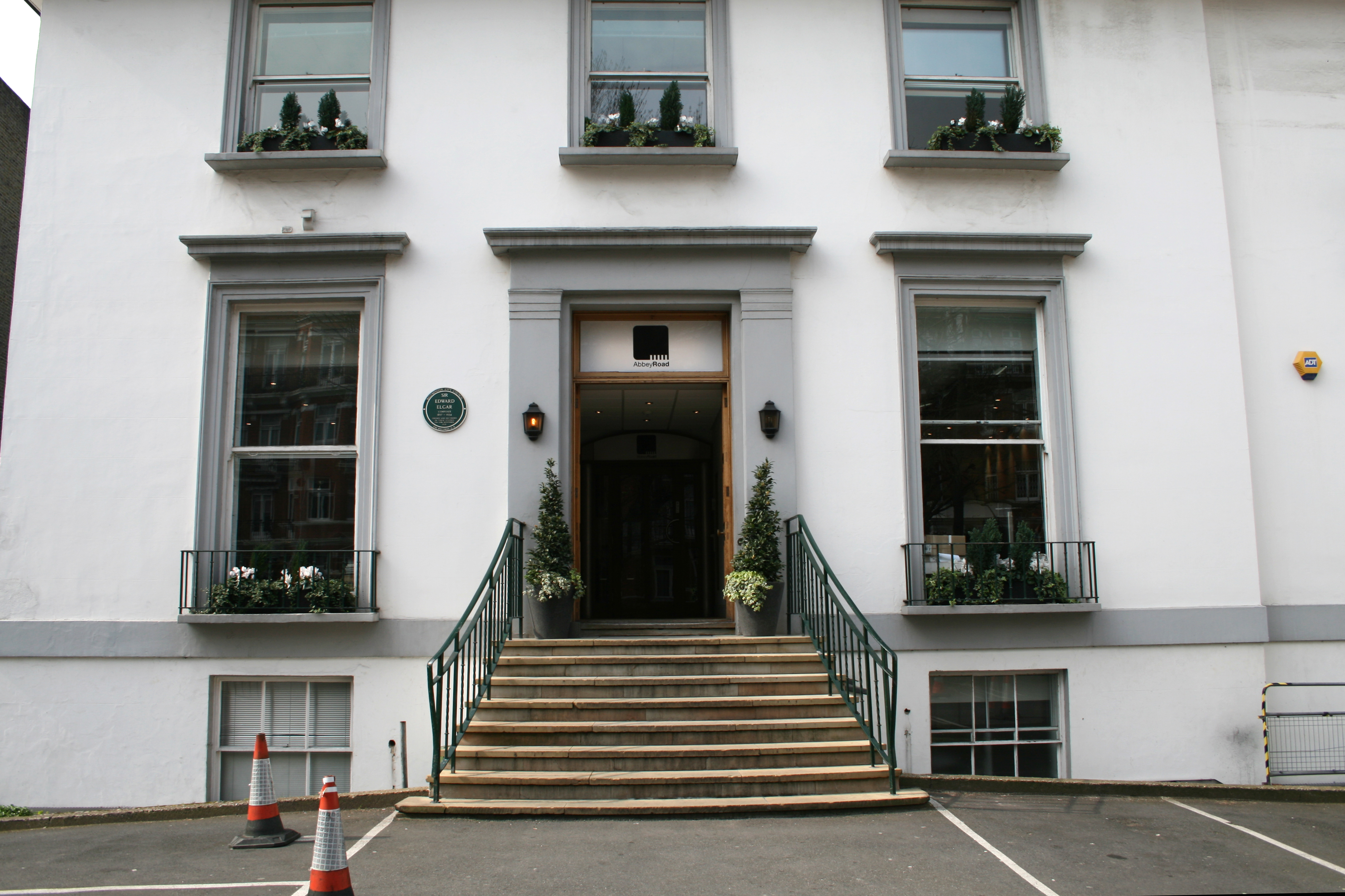 A flight of stone steps leads from an asphalt car park up to the main entrance of a white two-story building. The ground floor has two sash windows, the first floor has three shorter sash windows. Two more windows are visible at basement level. The decorative stonework around the doors and windows is painted grey.