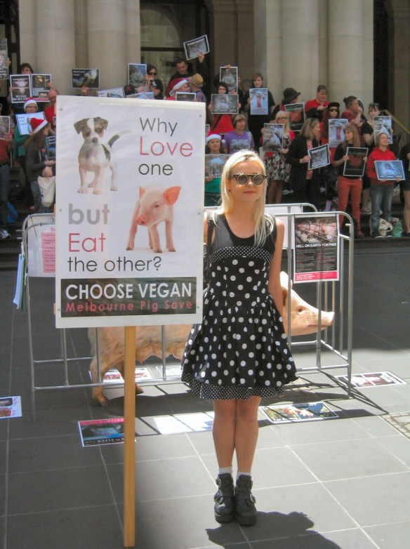 Woman with placard in front of 'pig' in cage, protesters in background - "Why Love One but Eat the Other?