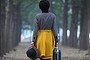 Woman in stylish clothes walks away with her back to the camera with a hat and small suitcase in her hands.