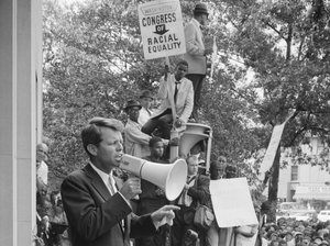 File - Attorney General Robert F. Kennedy speaking to a crowd of African Americans and whites through a megaphone outside the Justice Department; sign for Congress of Racial Equality is prominently displayed, 14 June, 1963.