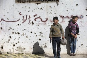 A Syrian boy holds a toy gun as he plays soccer with others between destroyed buildings with graffiti that reads "Syria al-Assad," in the old city of Homs, Syria, Friday, Feb. 26, 2016.