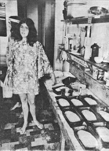 Photo of tired-looking young woman in kitchen surrounded by many loaves of baked bread.