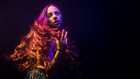 A woman with long hair stands against a black background, reaching out.